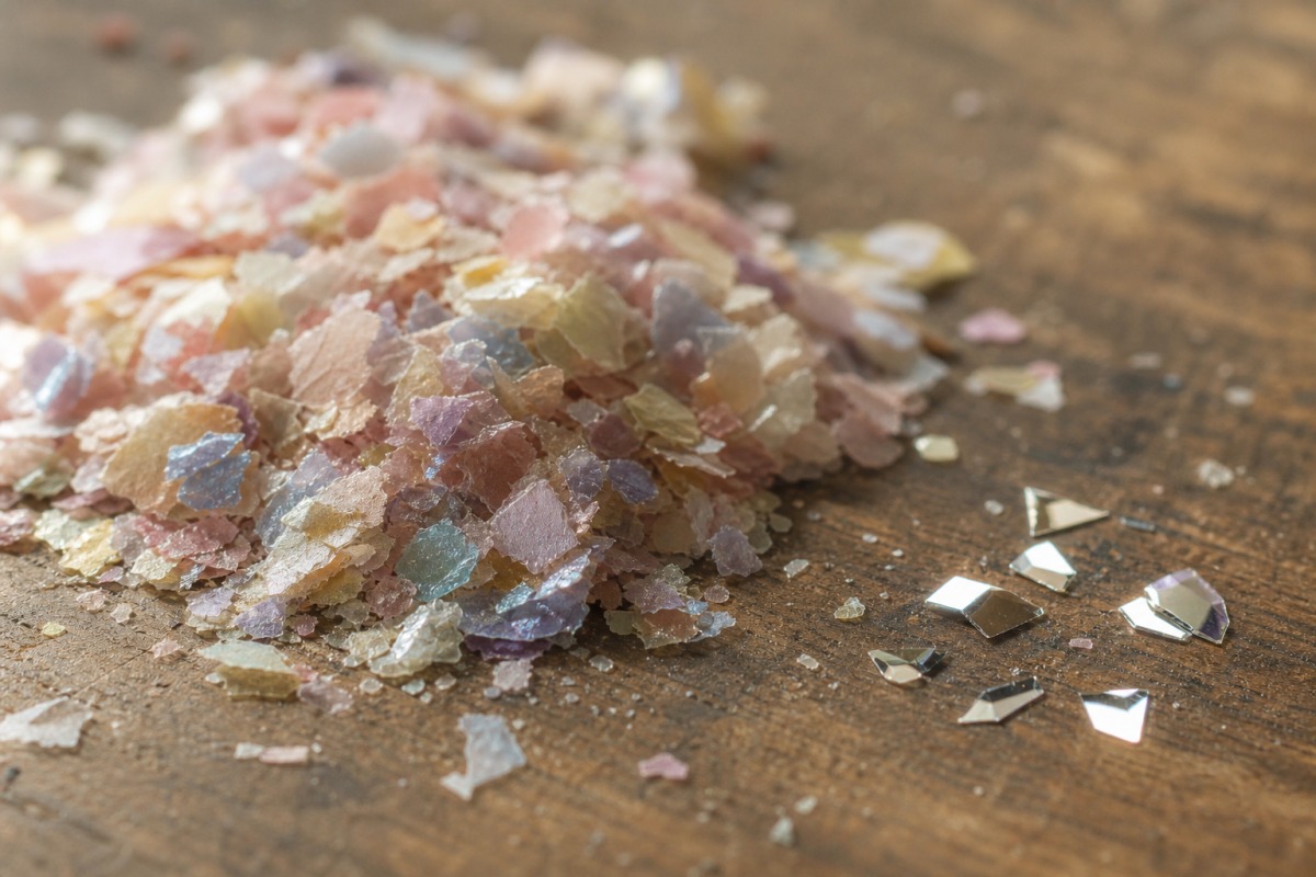 Dettaglio di un mucchietto di bio-glitter di cellulosa, più opaco, accanto ad alcune particelle di PET sintetico lucide su una superficie di legno.